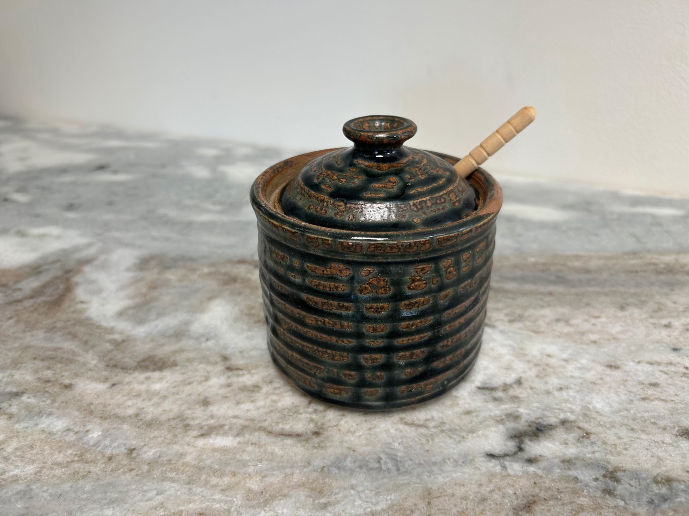 Handmade dark green stoneware honey pot with carved texture, fitted lid, and wooden honey dipper