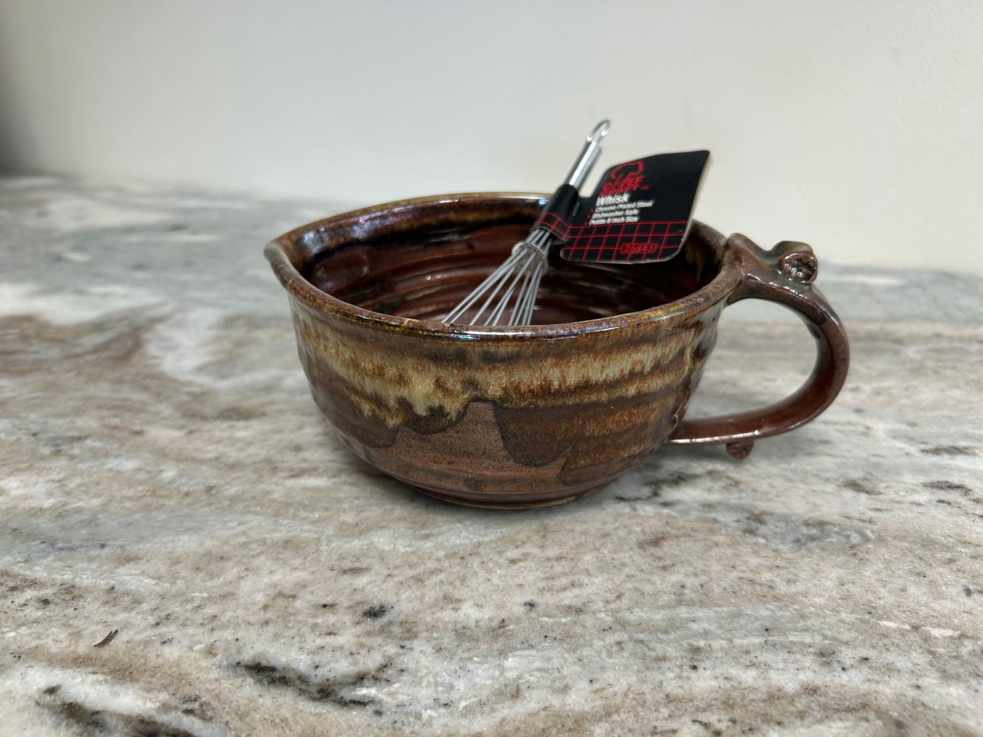Handmade pottery mixing bowl with rust body, cream drip rim, loop handle, and steel whisk