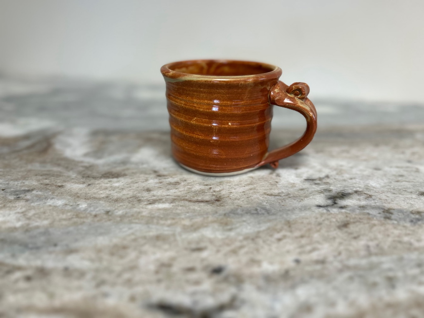 Small handmade pottery cup in glossy rust-orange glaze with curled handle