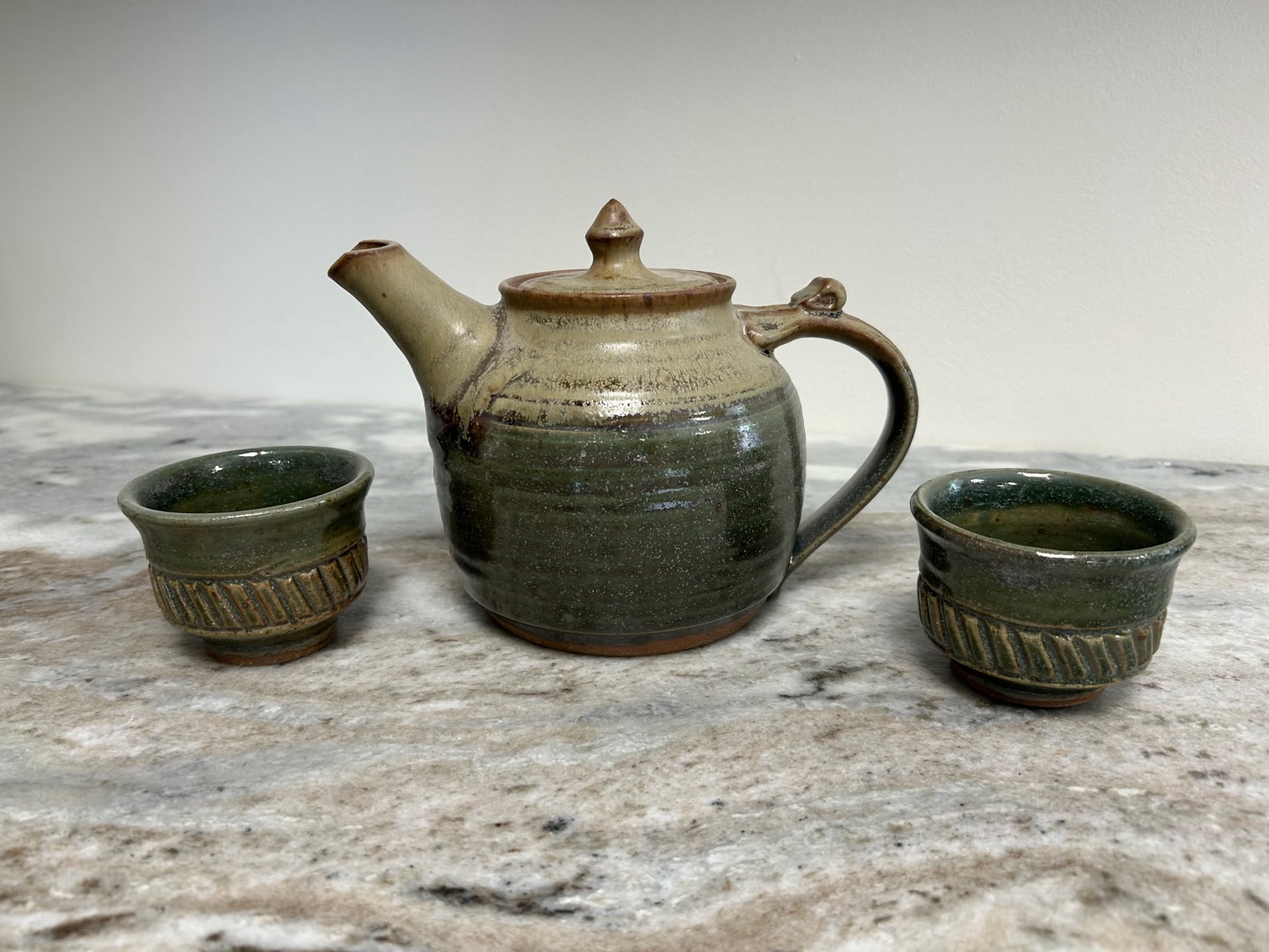 Handmade sage green pottery teapot with cone-finial lid and two matching cups with carved feet